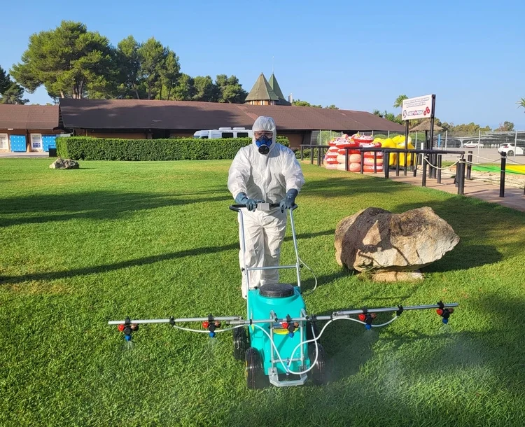Técnico con traje de protección y mascarilla aplicando tratamiento fitosanitario en césped con máquina pulverizadora