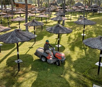 Un hombre vestido con una camiseta marrón y gorra verde opera una cortadora de césped montada en un extenso césped verde, rodeado de numerosas sombrillas de paja y árboles en un día soleado.